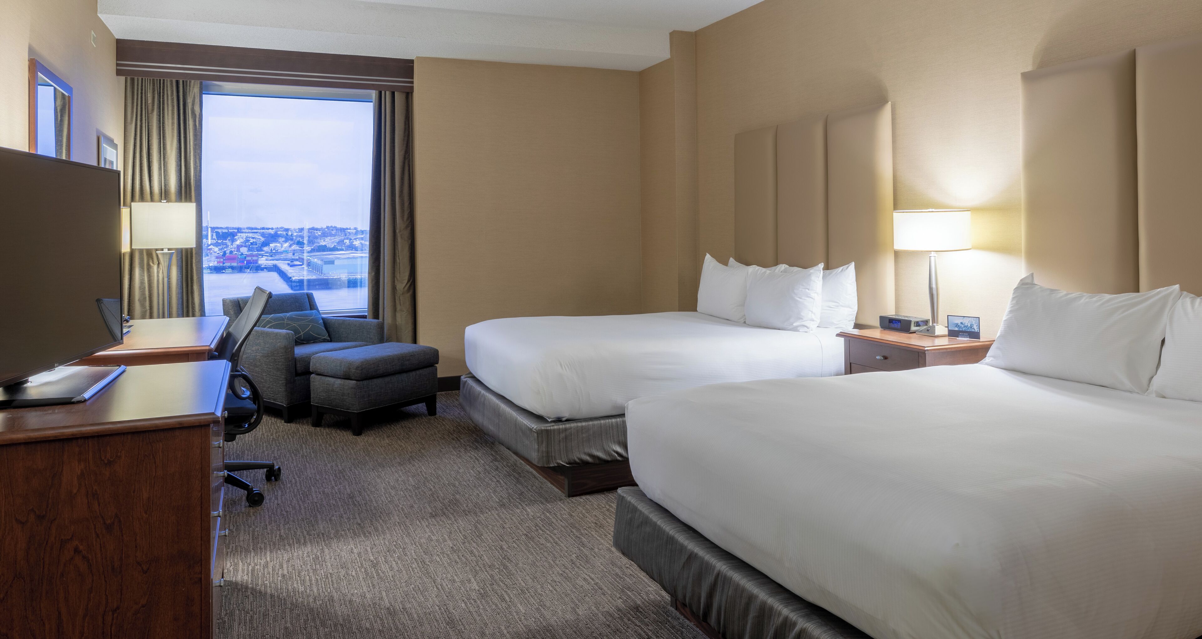 A hotel room with two beds, a desk, a chair, and a view of the city.