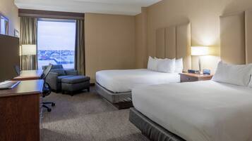 A hotel room with two beds, a desk, a chair, and a view of the city.