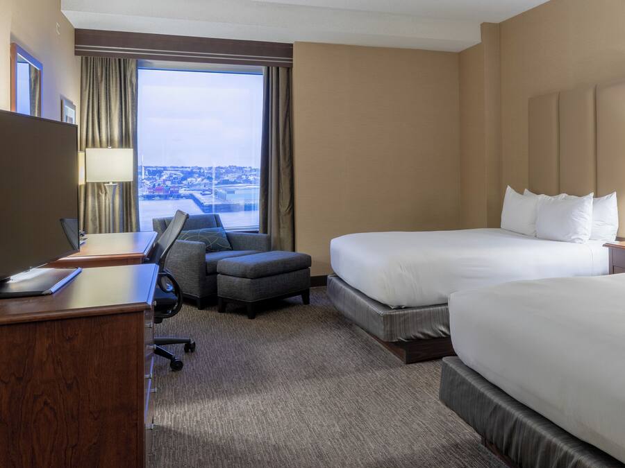 A hotel room with two beds, a desk, a chair, and a view of the city.