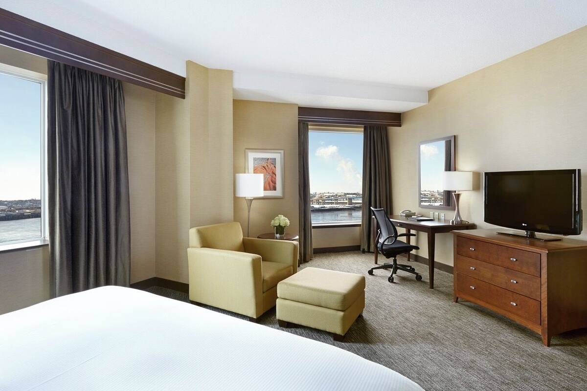 A hotel room with a large bed, a desk, a chair, a TV, and a view of the city.