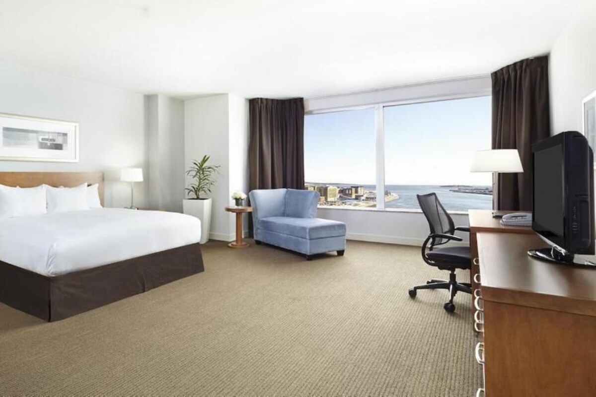 A hotel room with a large bed, a desk with a computer, a chair, a nightstand, and a window with a view.