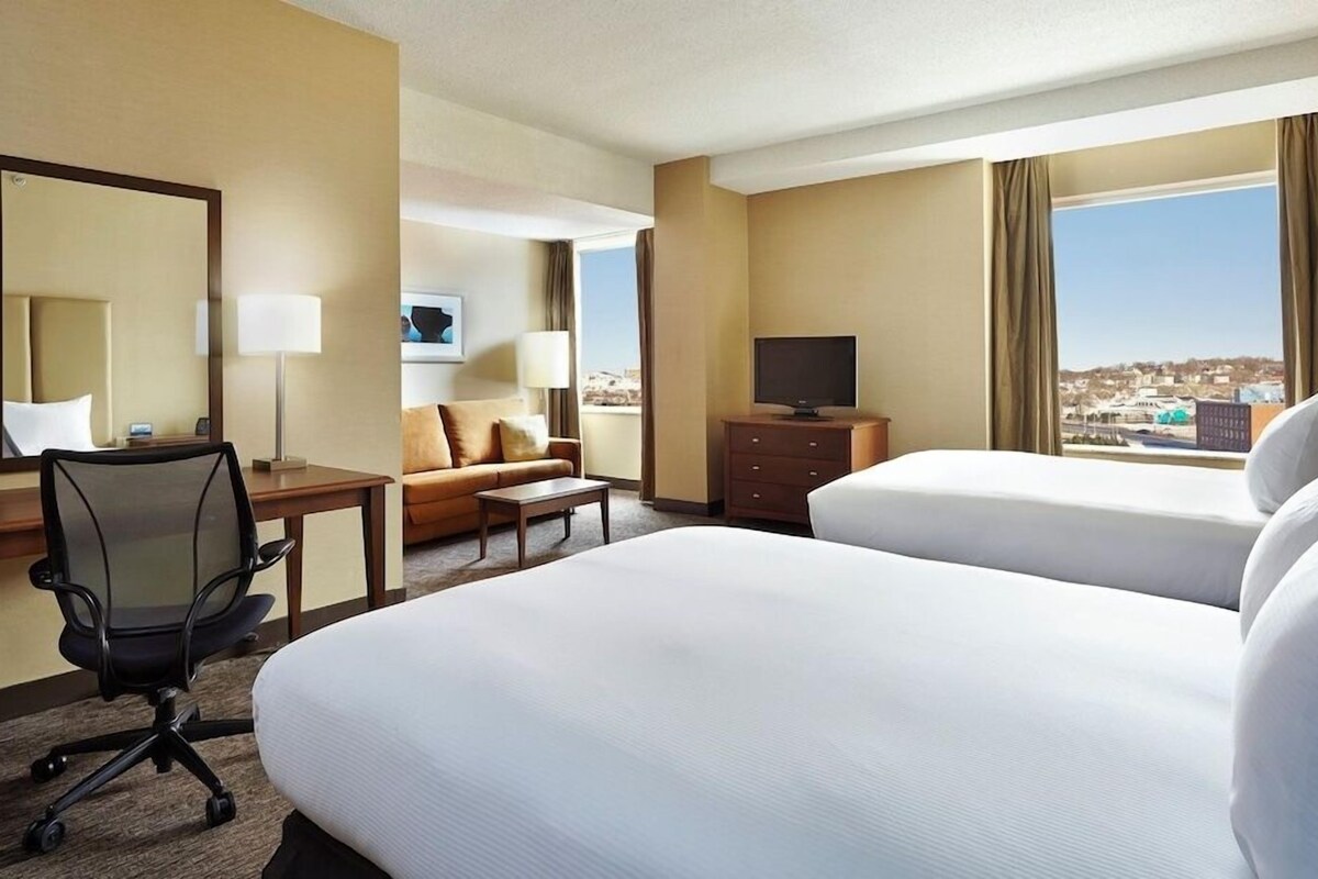 A hotel room with two beds, a desk with a chair, a television, and a view of the city.