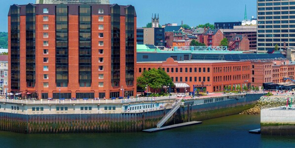 A waterfront building with a prominent red brick facade, a large glass window section, and a pedestrian bridge with stairs leading to the upper level. - Saint John Harbourfront Hotel (Saint John)