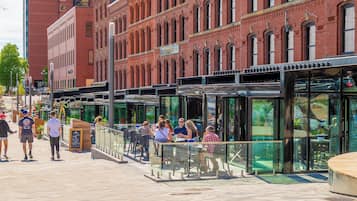 Un espace extérieur public aménagé avec des tables et des chaises, une structure fermée en verre et, en arrière-plan, un bâtiment en briques.