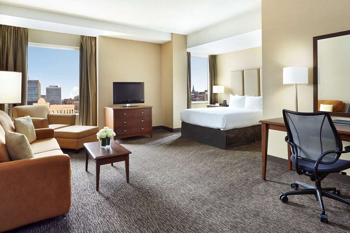 A hotel room with a bed, a desk, a chair, a TV, and a view of the city.