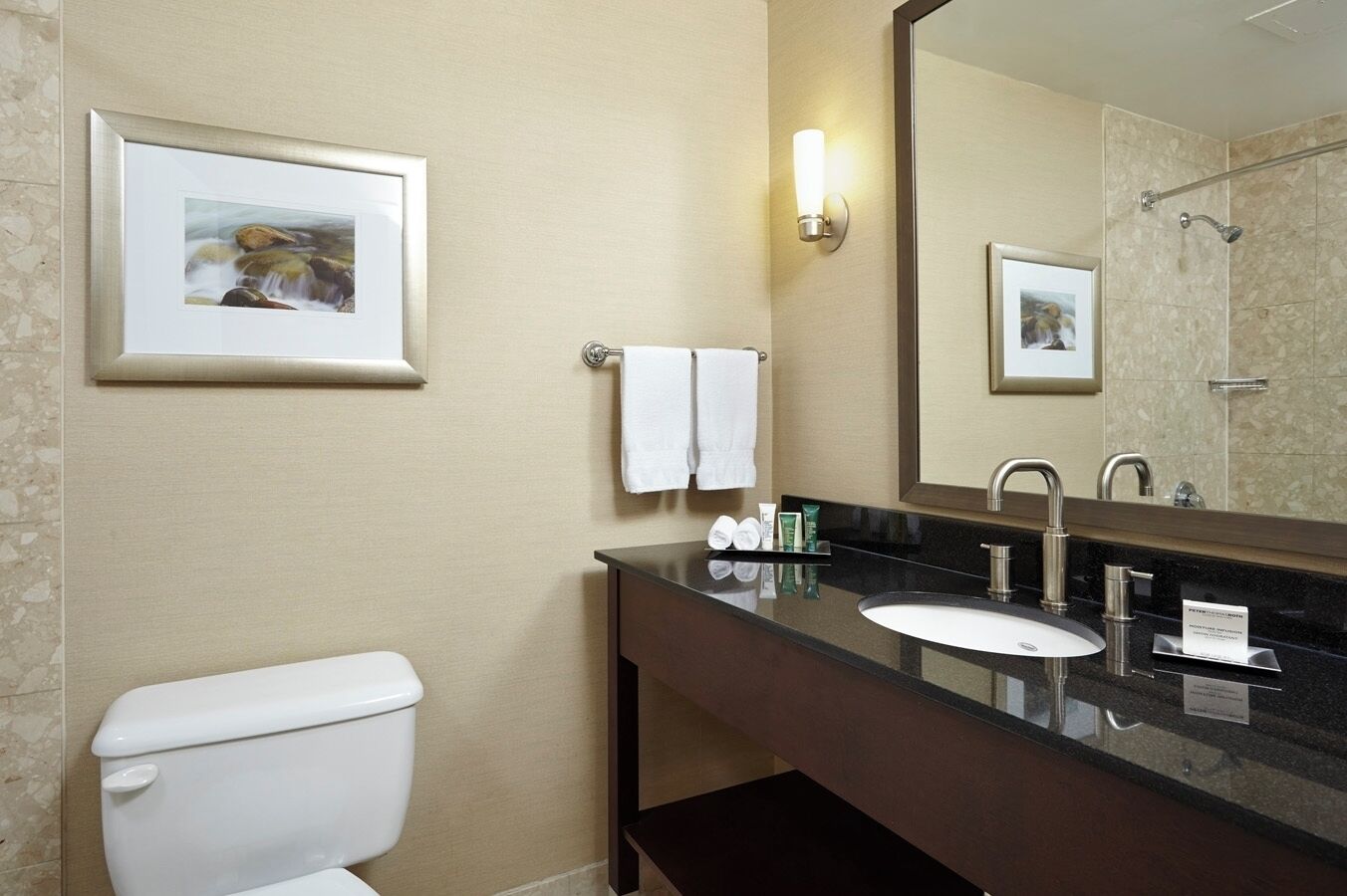 A modern bathroom with a toilet, sink, and mirror. There are towels hanging on a rack, a framed picture on the wall, and toiletries on the sink counter.