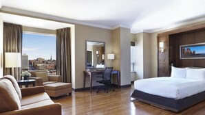 A hotel room with a bed, a desk, a chair, a sofa, and a large window with a city view.