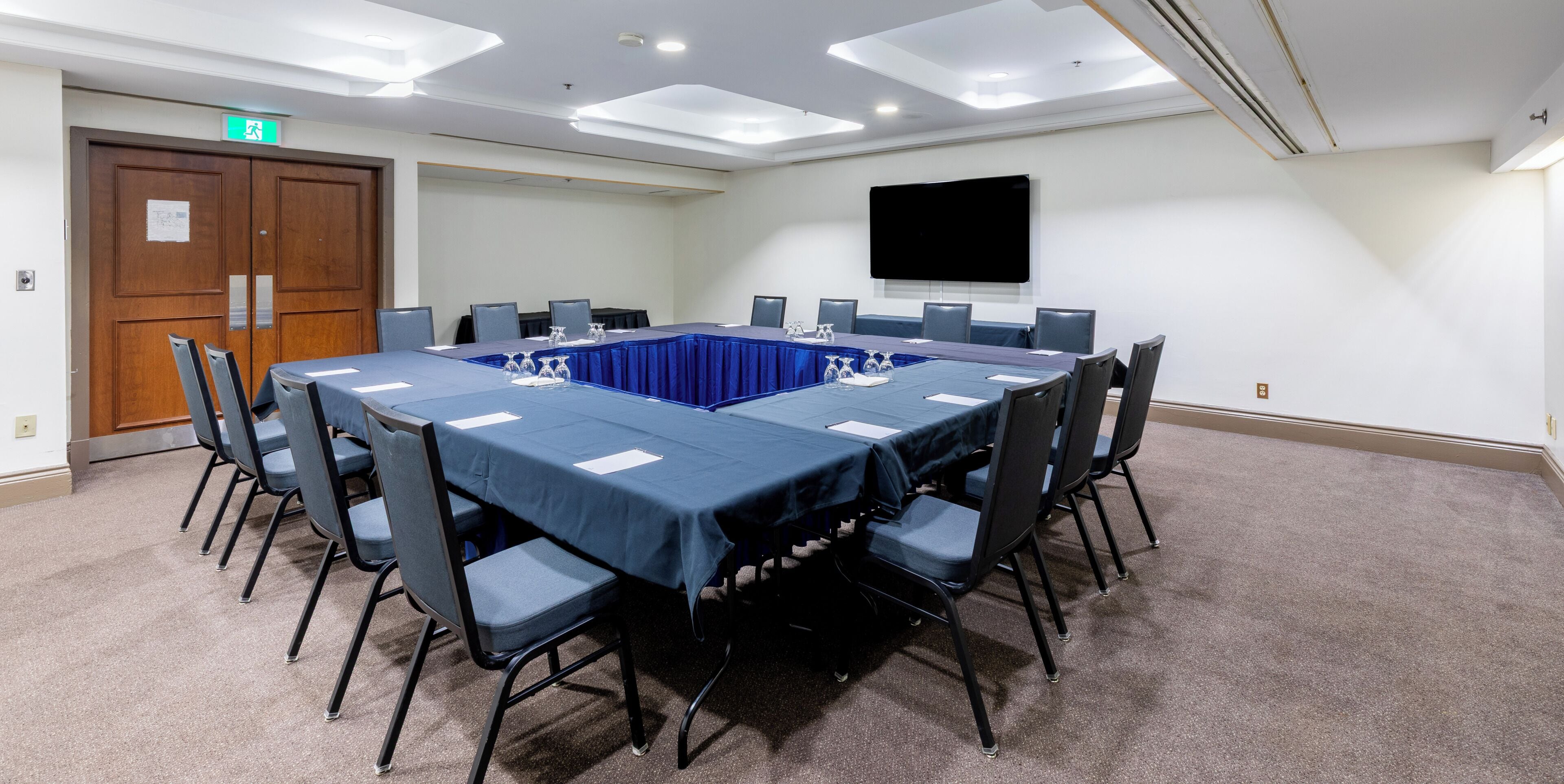 Un salón de conferencias con una larga mesa, sillas, un televisor de pantalla plana y una puerta con letrero de salida.