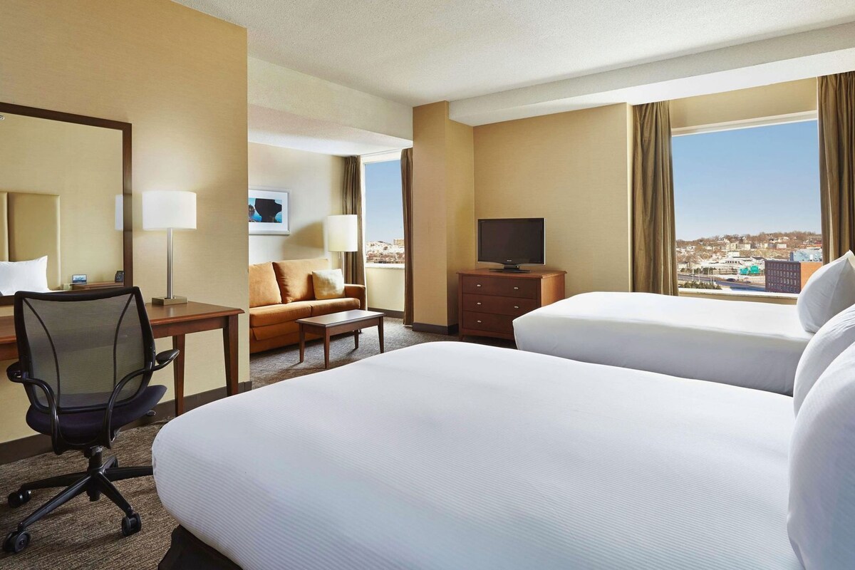 A hotel room with two beds, a desk, a chair, a TV, and a large window with a city view.