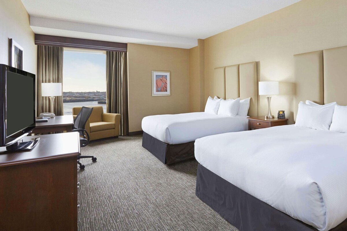 A hotel room with two beds, a desk with a TV, a chair, and a view of the city.