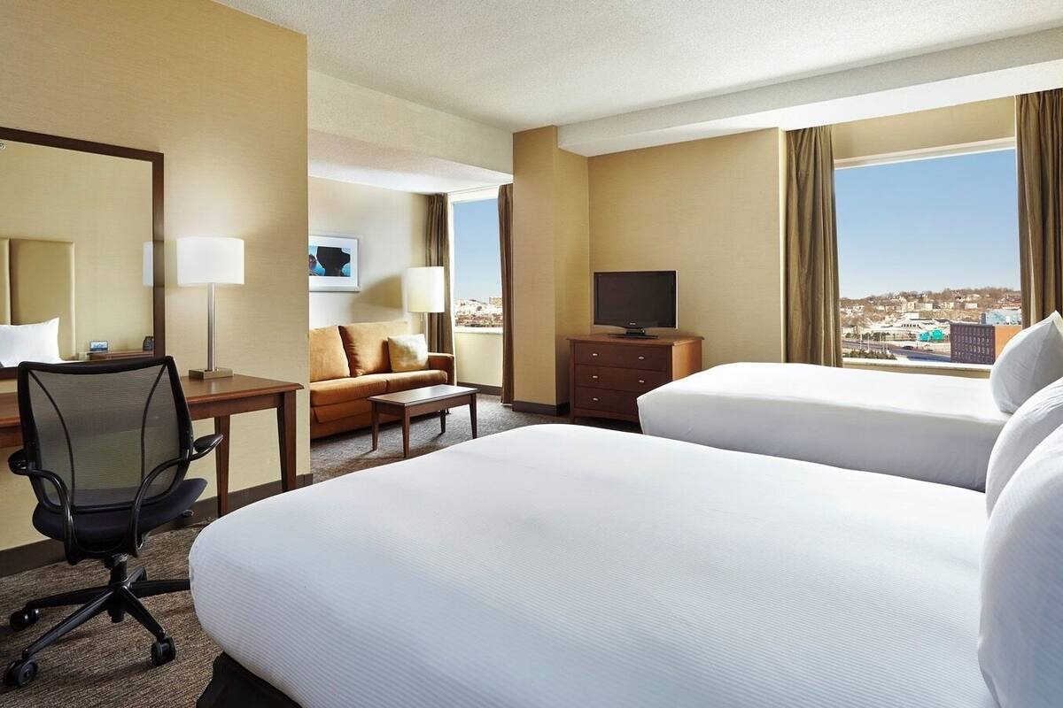 A hotel room with two beds, a desk with a chair, a TV, and a view of the city.