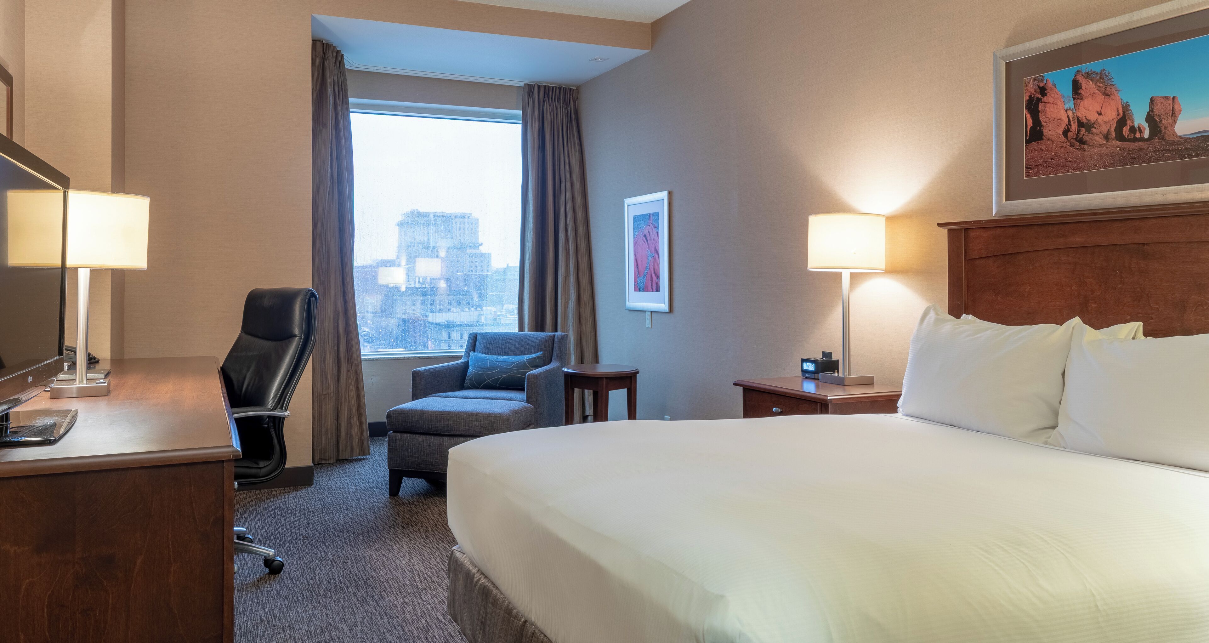 A hotel room with a bed, desk, chair, lamp, and a view of the city through the window.