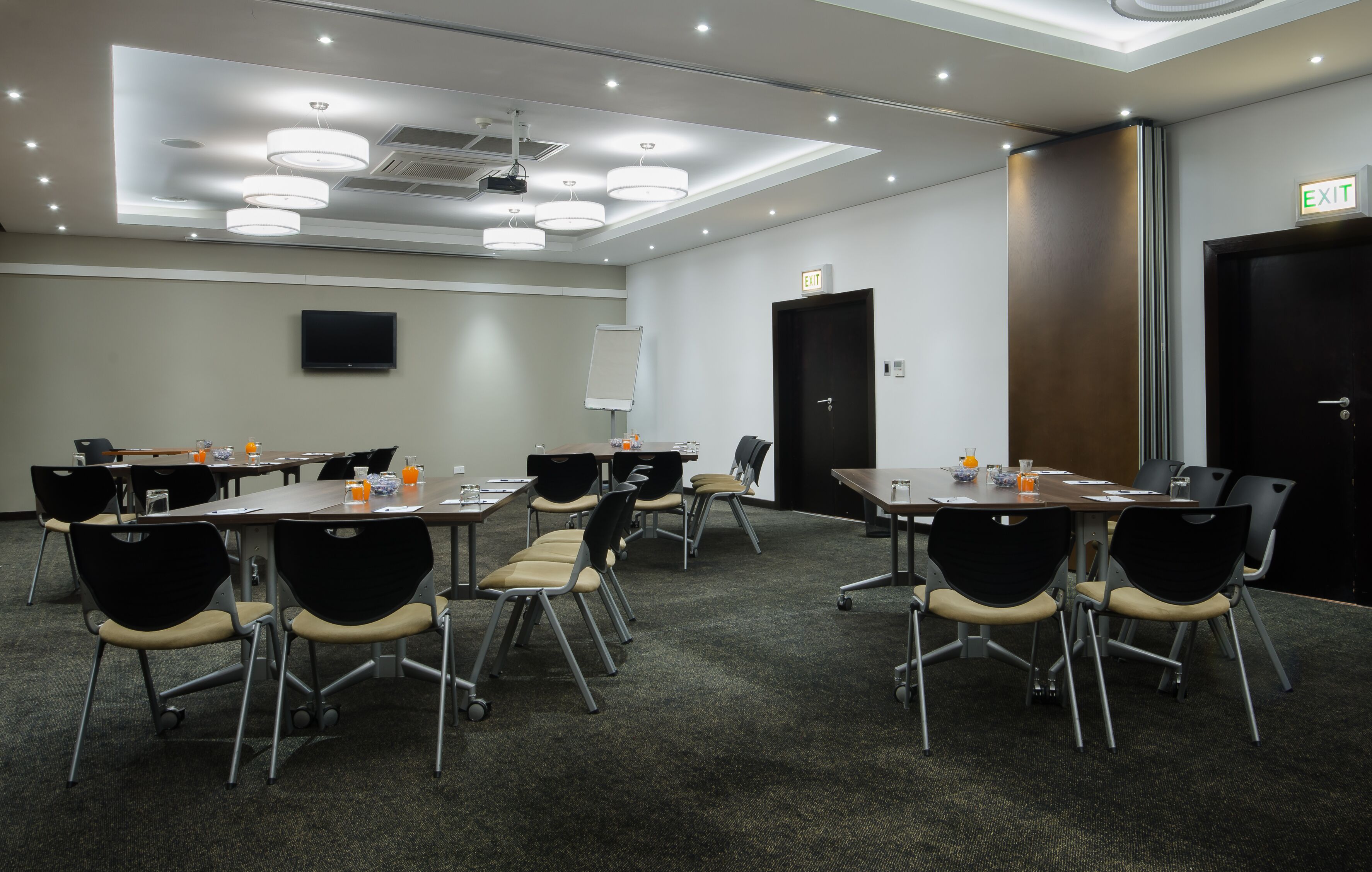 A conference room with tables, chairs, a whiteboard, a TV, and an exit sign.