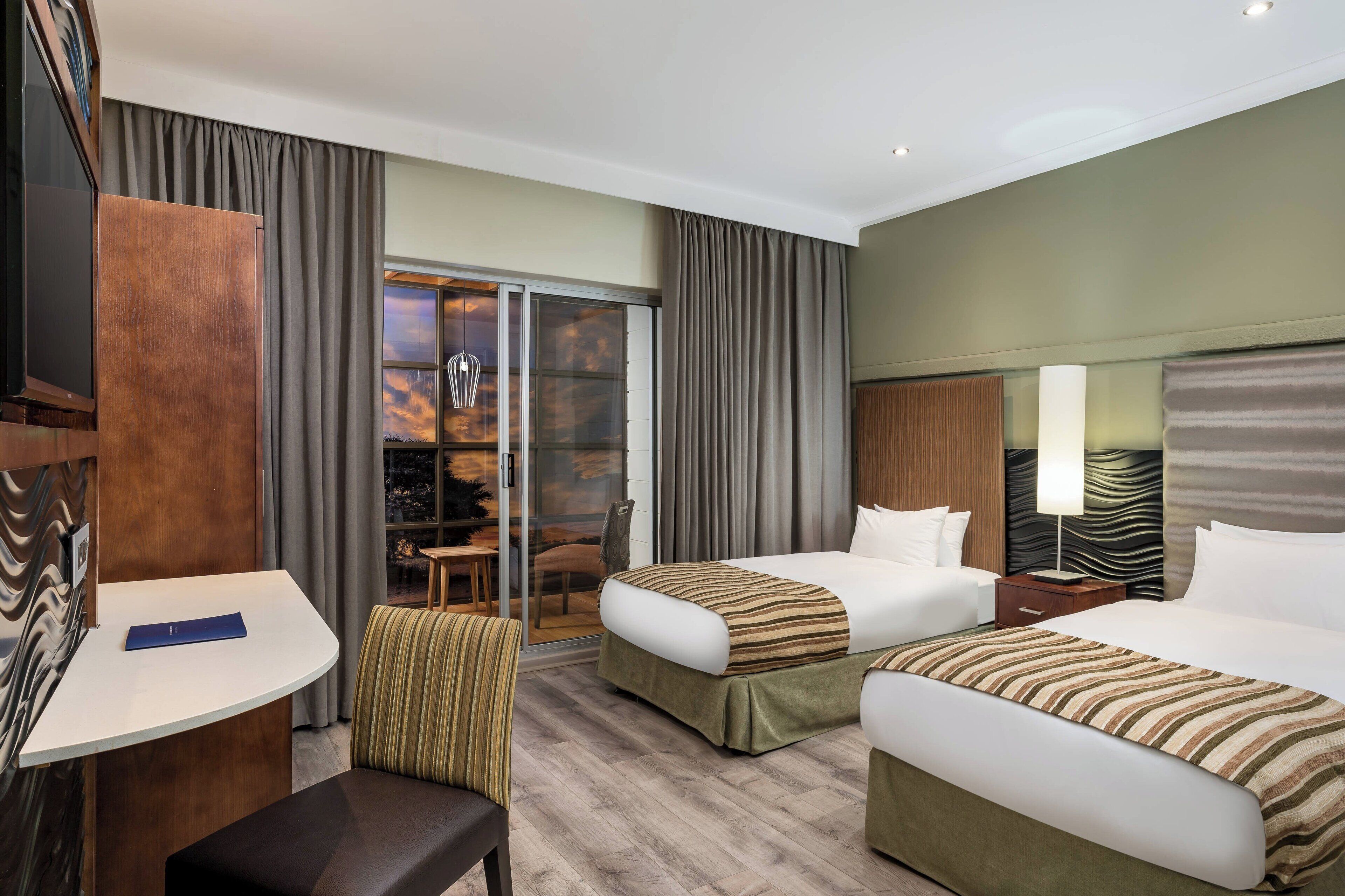 A hotel room with two beds, a desk, a chair, a TV, and a sliding glass door leading to a balcony.