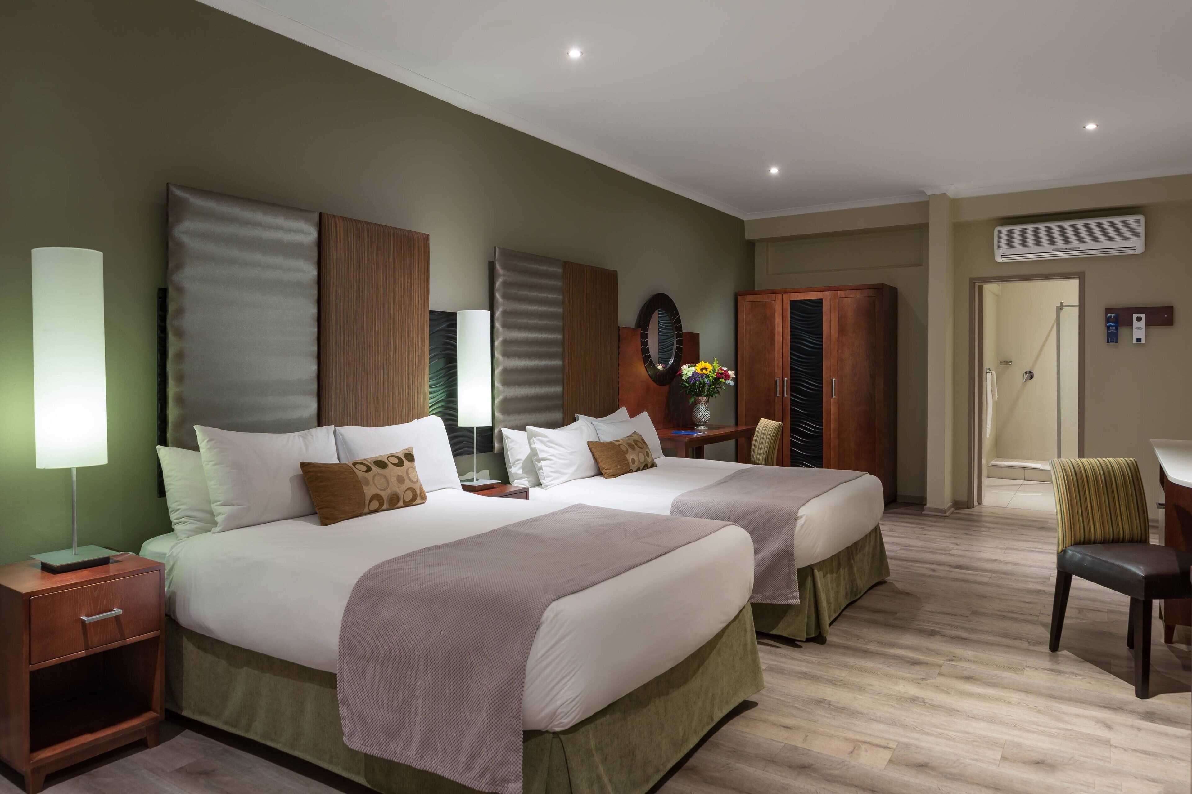 A hotel room with two beds, a nightstand, a chair, a desk, and a mirror.