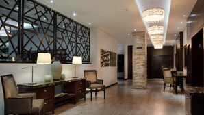Un hall d’hôtel spacieux au design moderne, comprenant un bureau en bois, deux chaises, un panneau mural décoratif et un grand lustre.