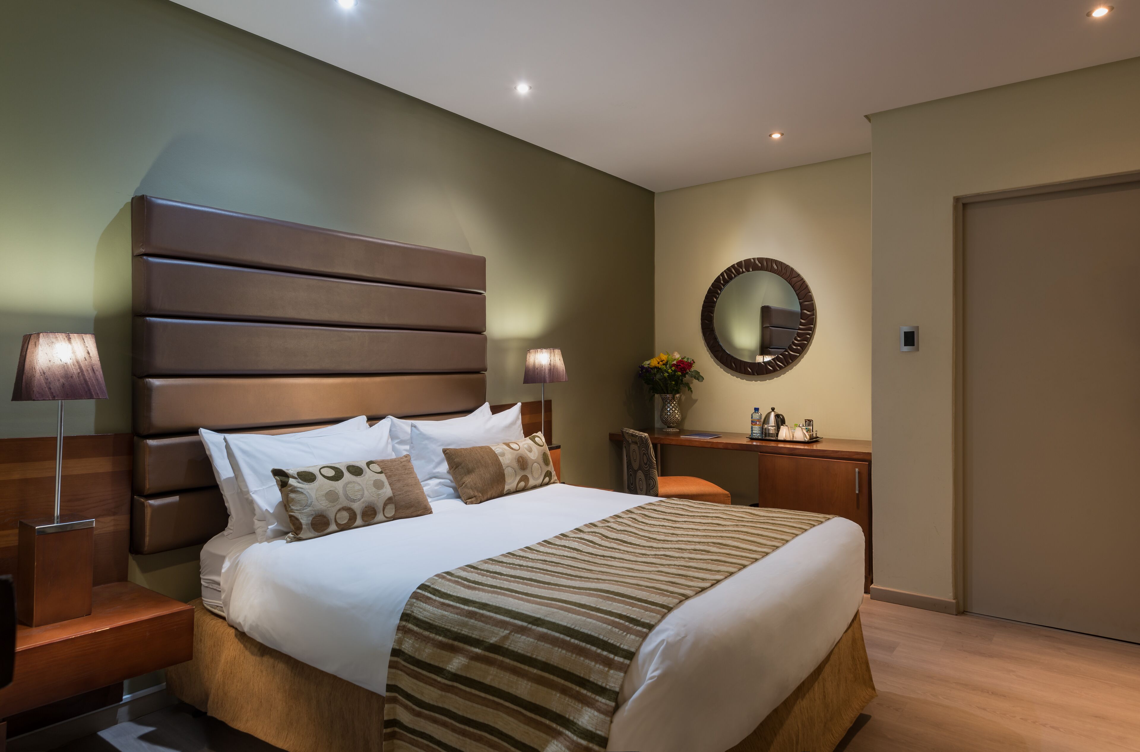 Ett modernt hotellrum med en stor säng, ett träbeklätt sänggavel, sänglampor, en rund spegel och ett litet skrivbord med en vas med blommor.