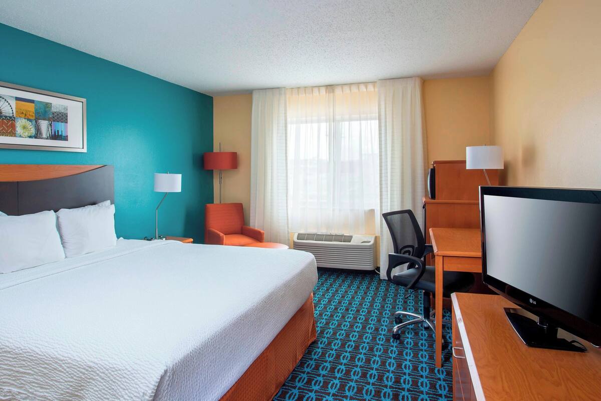 A hotel room with a large bed, a desk with a computer, a chair, an air conditioning unit, and a television.