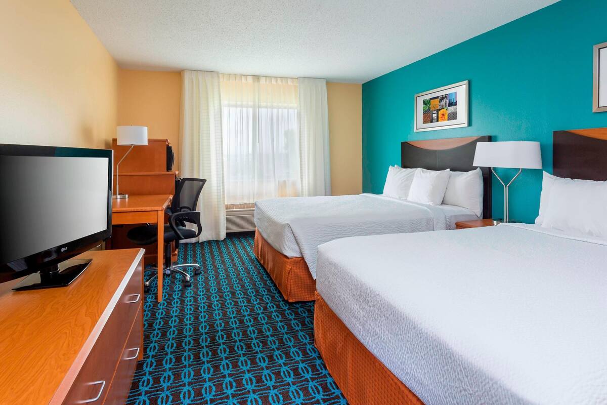 A hotel room with two beds, a desk with a TV, a chair, a desk lamp, and a window with curtains.