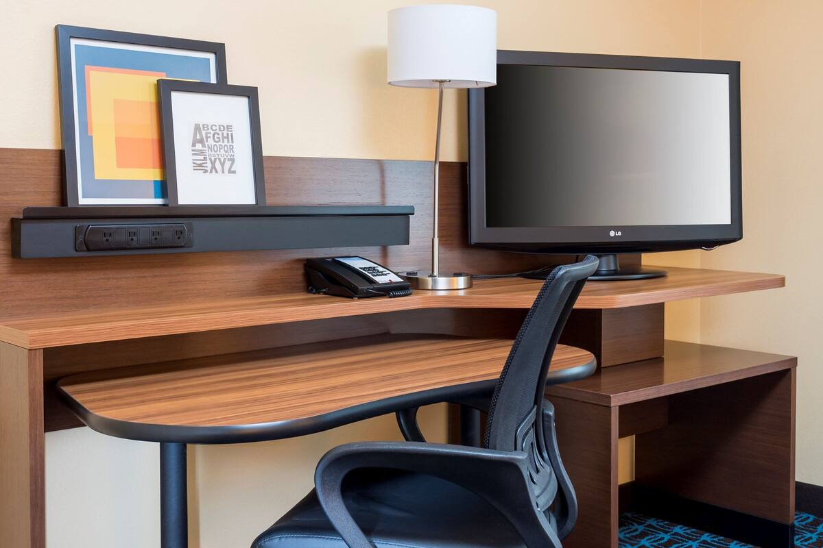 A hotel room desk with a flat-screen TV, a lamp, a phone, and a framed picture on the wall.
