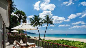 A balcony with a lounge chair and a table, overlooking a beach with palm trees and a sailboat.
