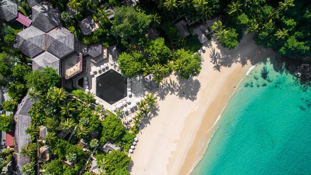 An aerial view of a beach resort with a hexagonal building surrounded by palm trees and a sandy beach.