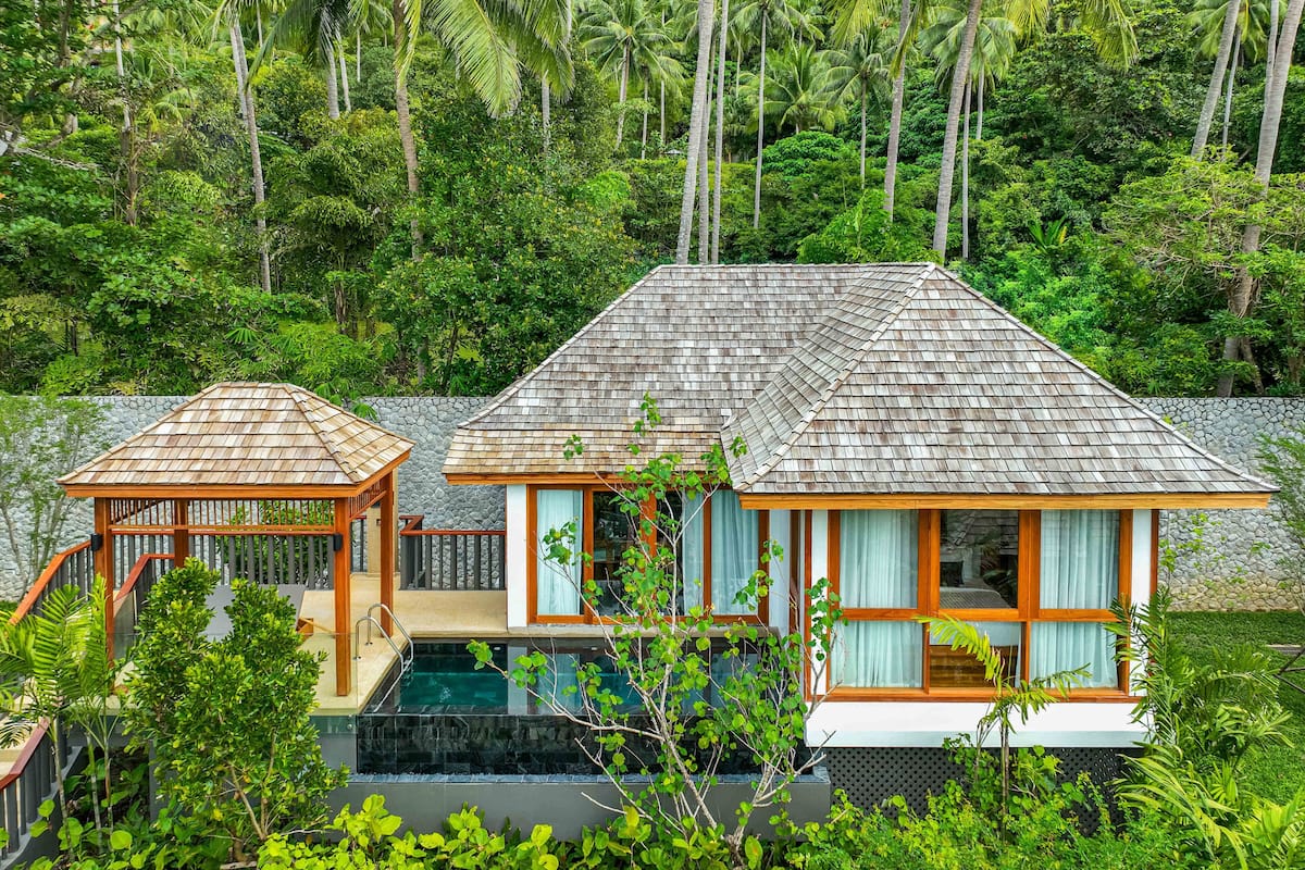 A two-story house with a swimming pool, surrounded by lush greenery and palm trees.
