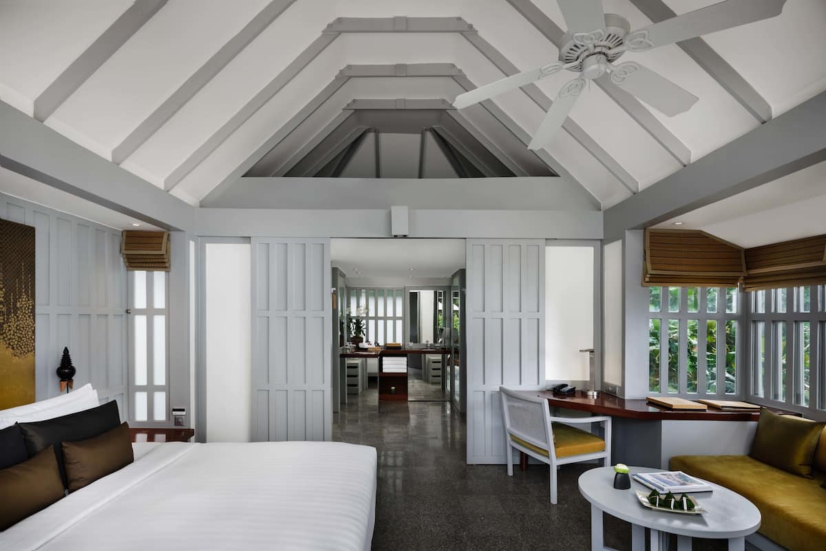 A modern hotel room with a large bed, a desk, a sofa, and a view of greenery through the windows.