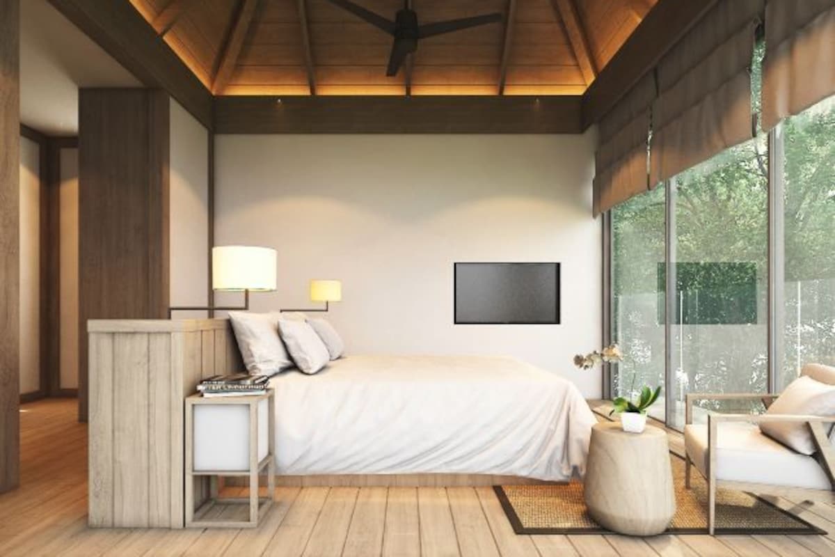 A modern bedroom with a large bed, bedside lamps, a wooden nightstand, a round side table, and a view of greenery through large windows.