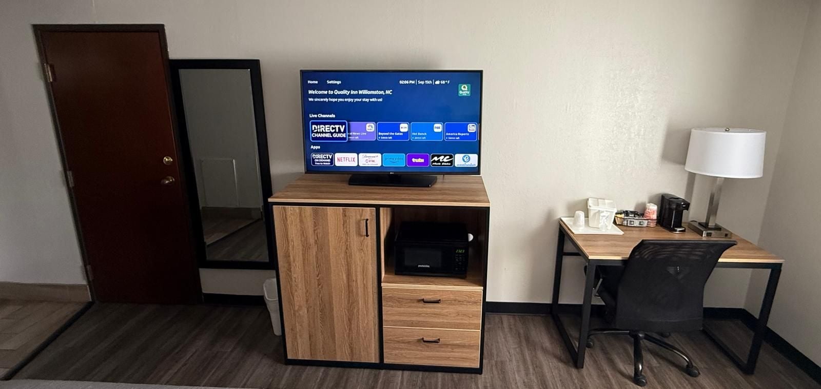 A hotel room with a wooden desk, a television displaying a channel guide, a microwave, and a coffee maker.