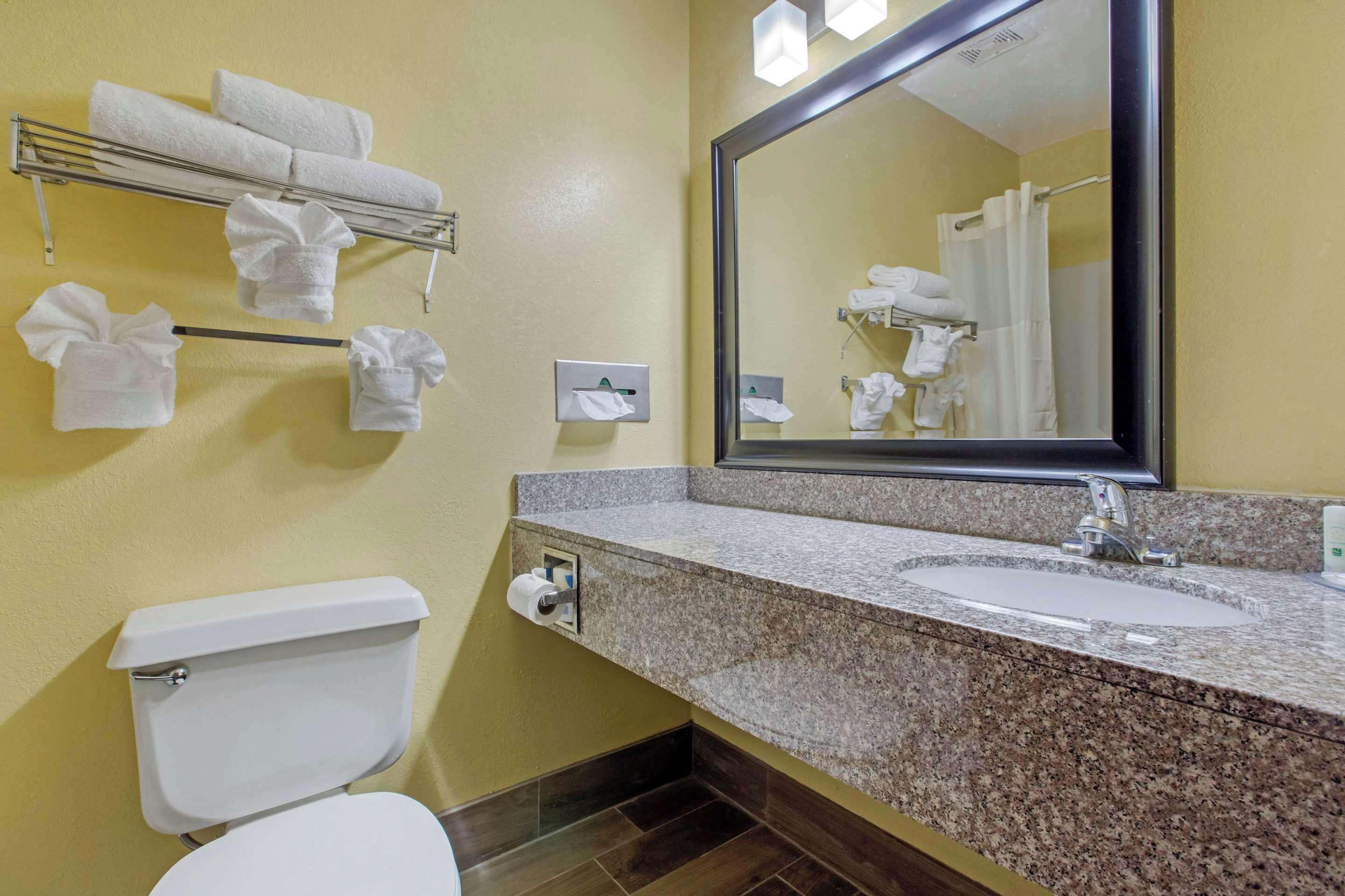 A bathroom with a toilet, sink, mirror, towel rack, and a towel dispenser.