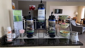 Zona de bar en un hotel con varios dispensadores de agua, un dispensador de jugo fresco y una variedad de cuencos y vasos sobre una barra.