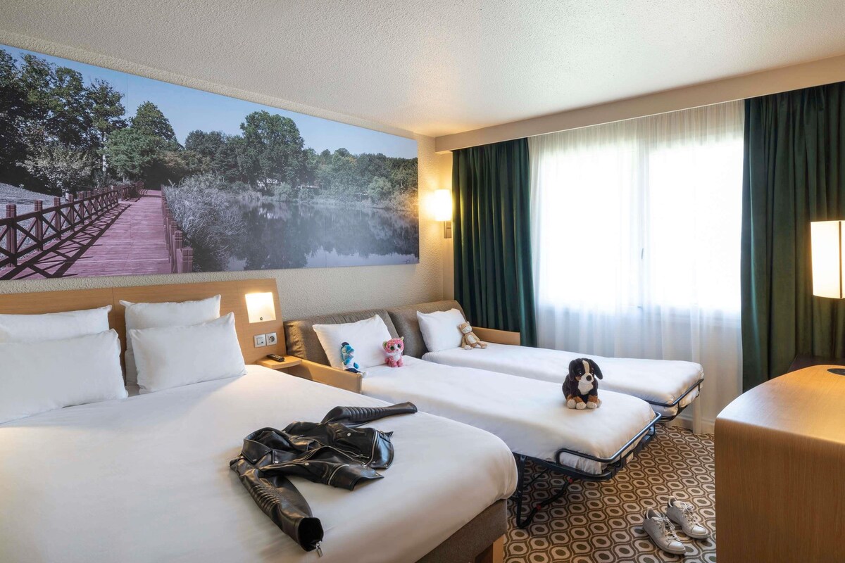A hotel room with two beds, a large mural of a bridge over water, a window with curtains, and a small table with a lamp.