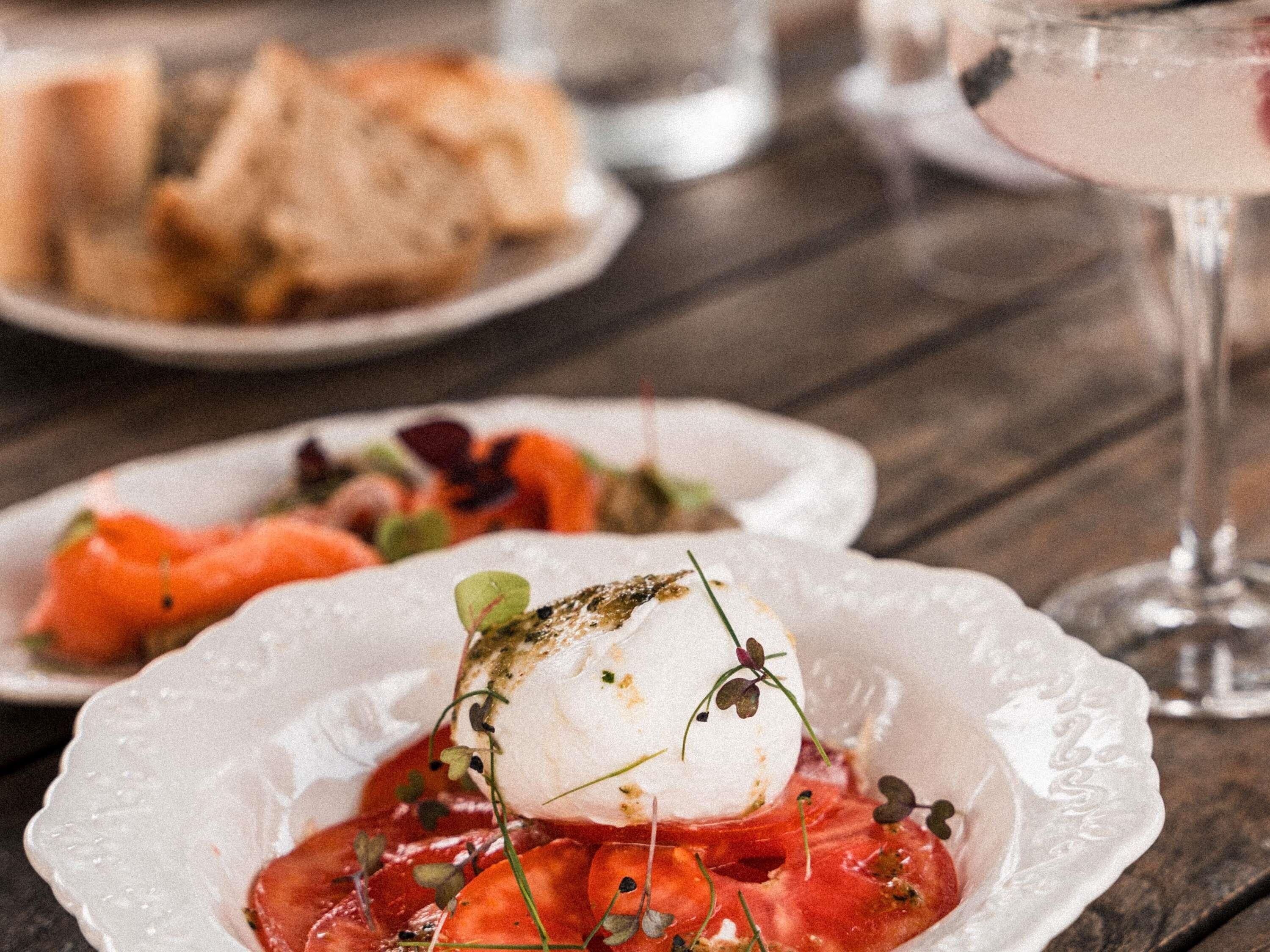 Een bord met een witte voedingsmiddel, mogelijk mozzarella, gegarneerd met groenten en tomaten, en een glas roze drankje op de achtergrond.