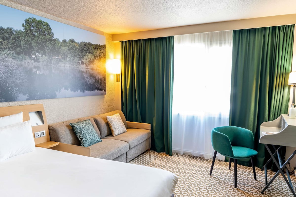 Habitación de hotel con una cama, un sofá, un escritorio y una silla. Hay un cuadro en la pared y una ventana con cortinas.
