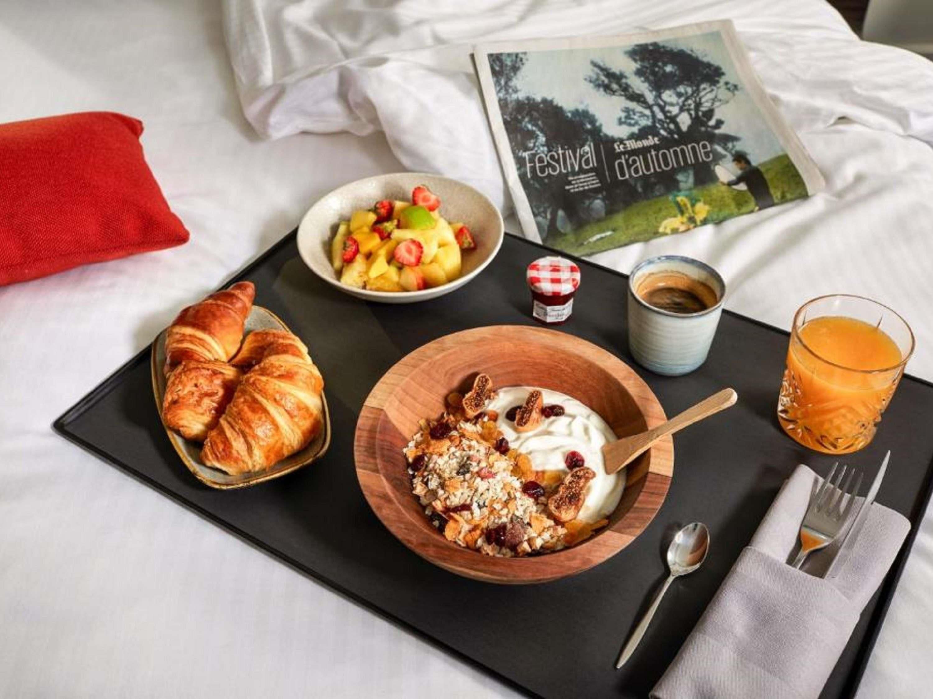 A breakfast tray with croissants, yogurt with fruit and granola, a glass of orange juice, and a magazine on a bed.