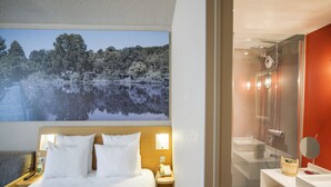 Habitación de hotel con cama, mesita de noche con lámpara, un cuadro de un lago y zona de ducha con espejo.