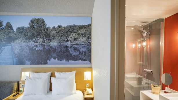 Habitación de hotel con cama, mesita de noche con lámpara, un cuadro de un lago y zona de ducha con espejo.
