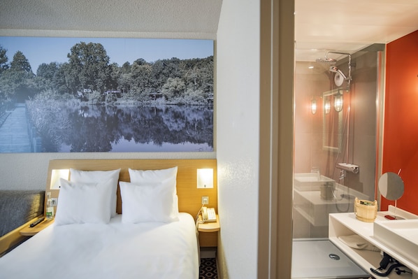 A hotel room with a bed, bedside table with a lamp, a painting of a lake, and a shower area with a mirror.