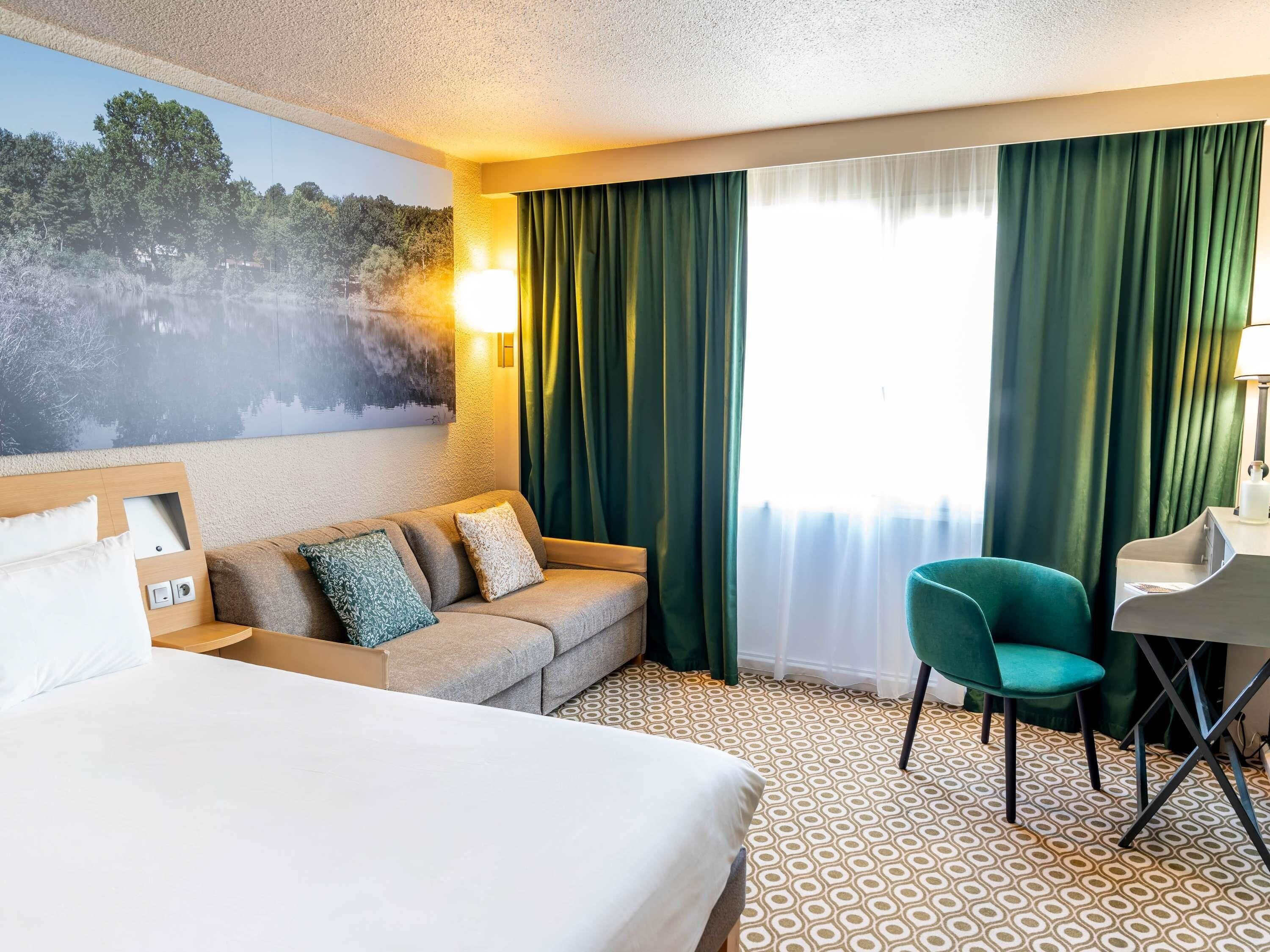 Une chambre d’hôtel avec un lit, un canapé, un bureau et une chaise. Il y a un tableau au mur et une fenêtre avec des rideaux.