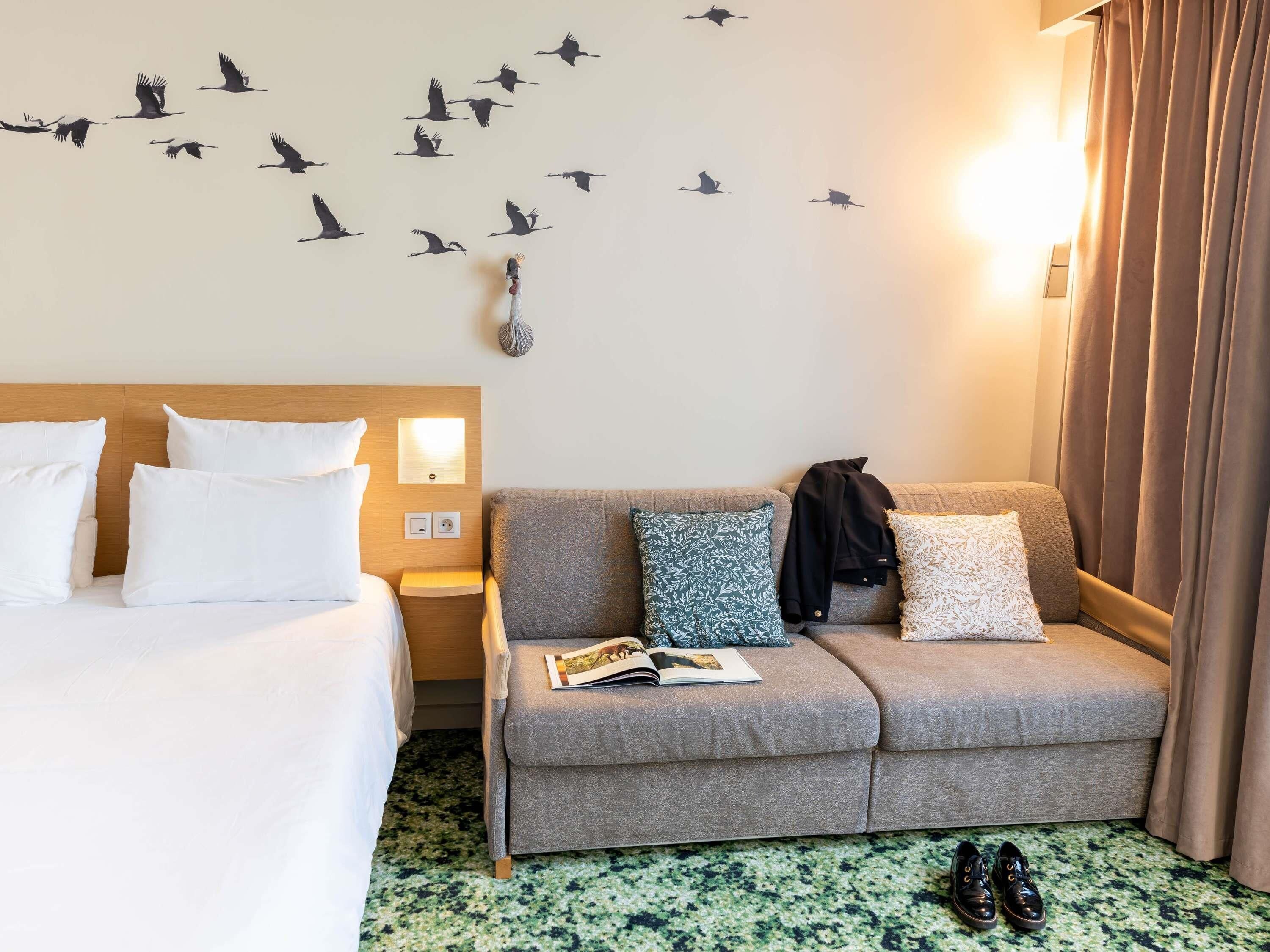 A hotel room with a bed, a sofa, a wall-mounted light, a wall decoration of birds, a nightstand, and a pair of shoes on the floor.