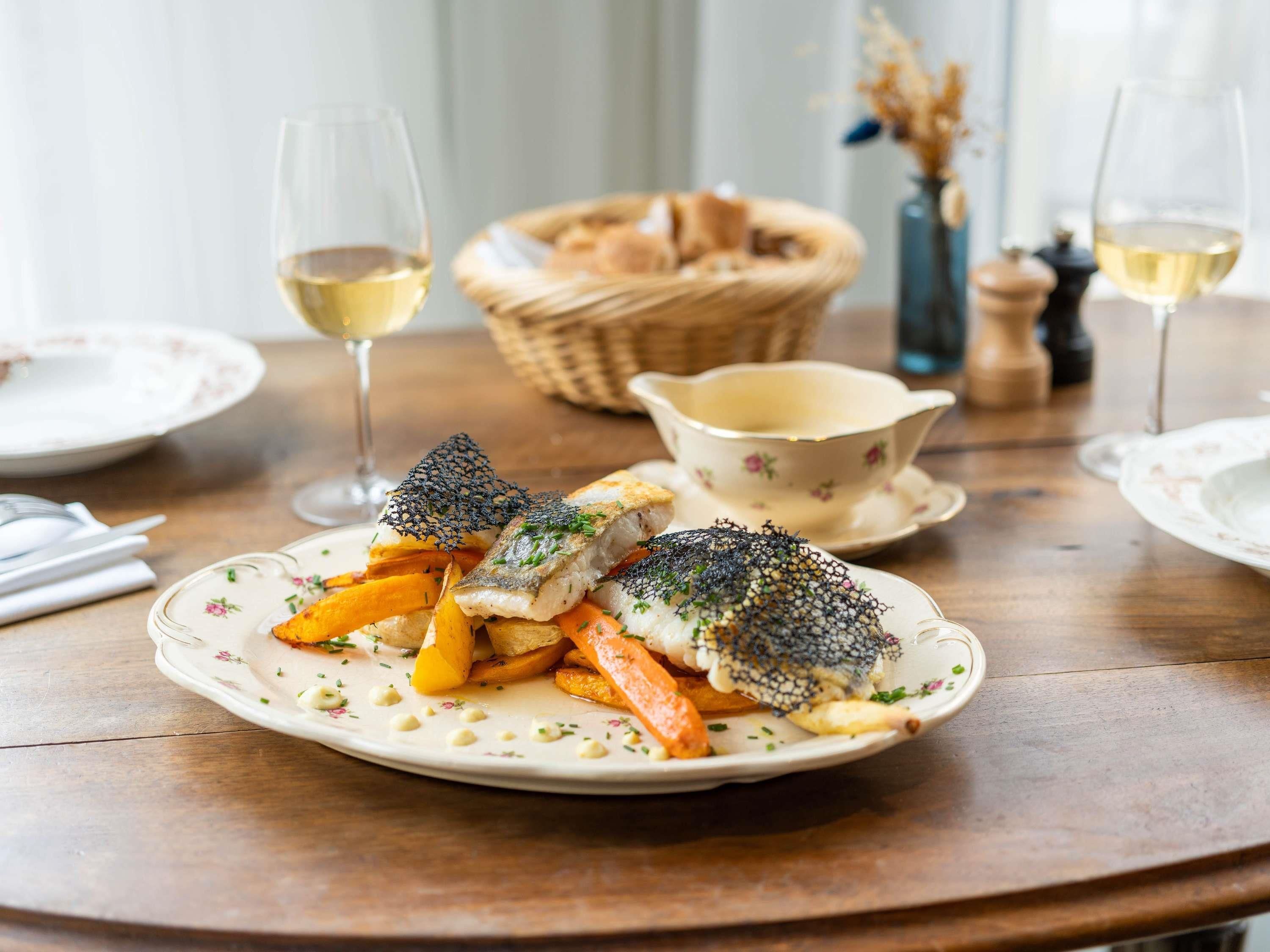 A dining table with a plate of fish, fries, and a decorative net on top. There are glasses of white wine and a basket of bread in the background.