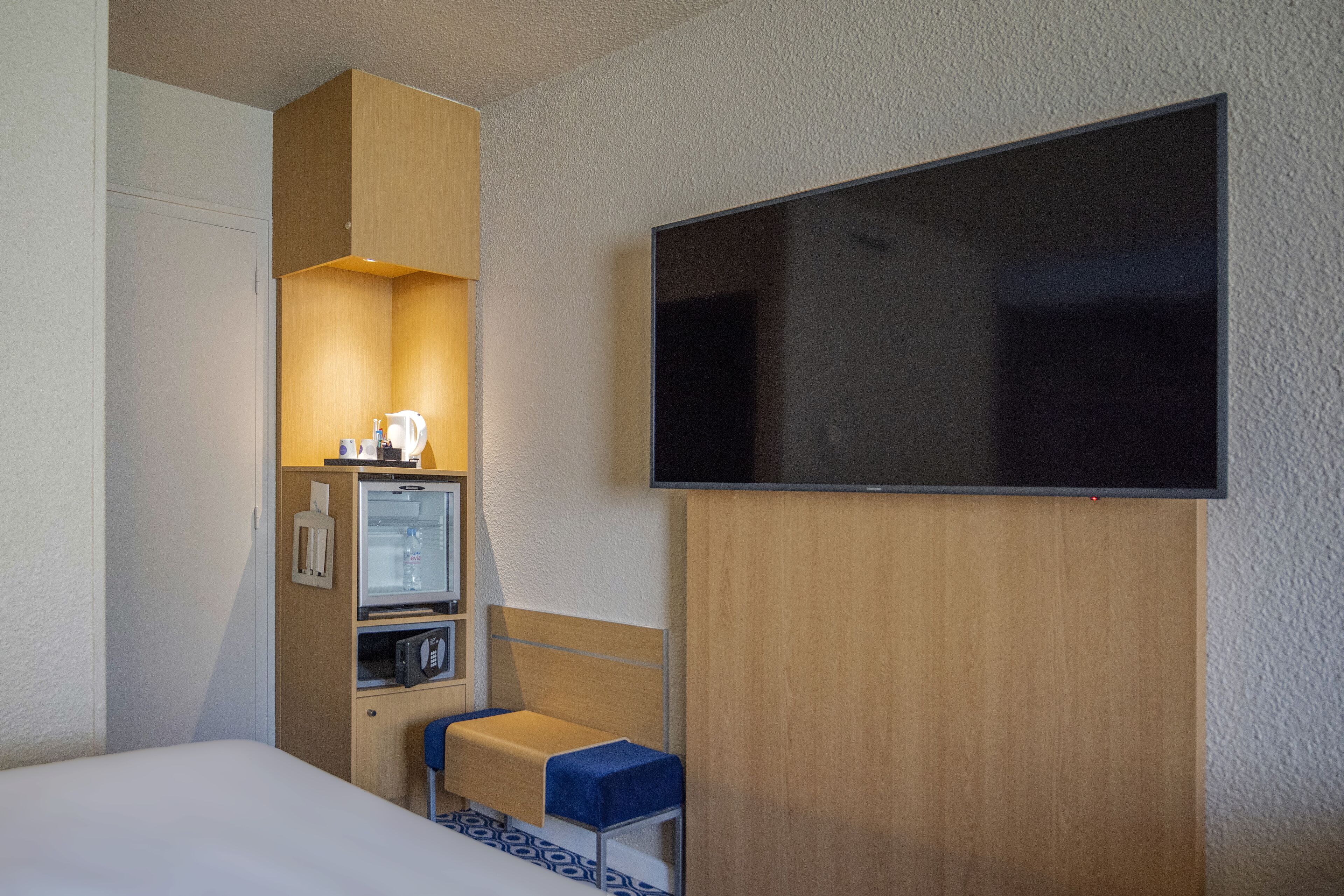 Ein Hotelzimmer mit Bett, Wandfernseher, kleinem Schreibtisch mit Stuhl und Minibar.