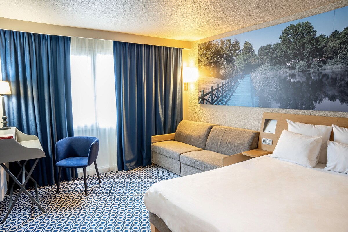 A hotel room with a bed, a sofa, a desk, and a chair. There is a painting of a bridge over water on the wall.