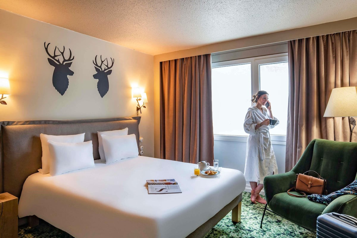 Una habitación de hotel con una cama grande, un sillón verde, una mesita de noche y un ventanal con cortinas.