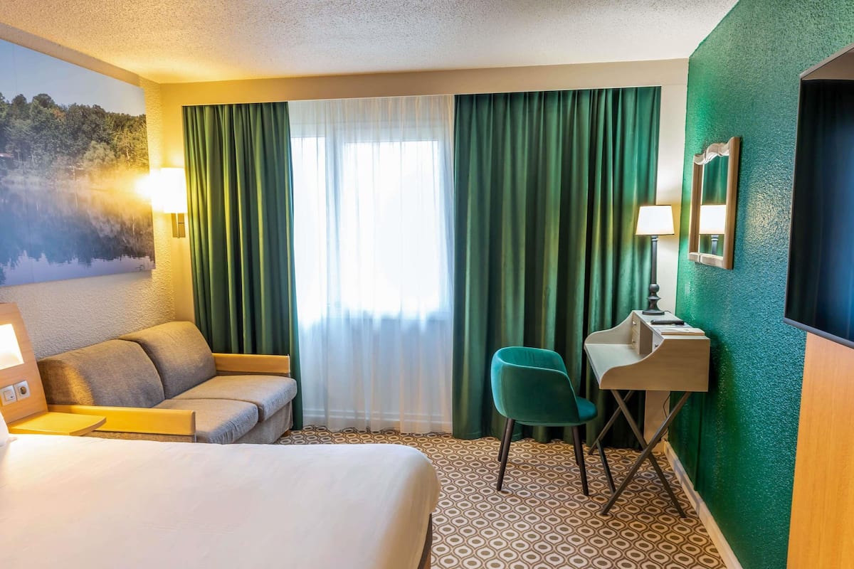 Ein Hotelzimmer mit einem Bett, einem Schreibtisch mit Stuhl, einem großen Fenster mit Vorhängen und einem an der Wand montierten Fernseher.