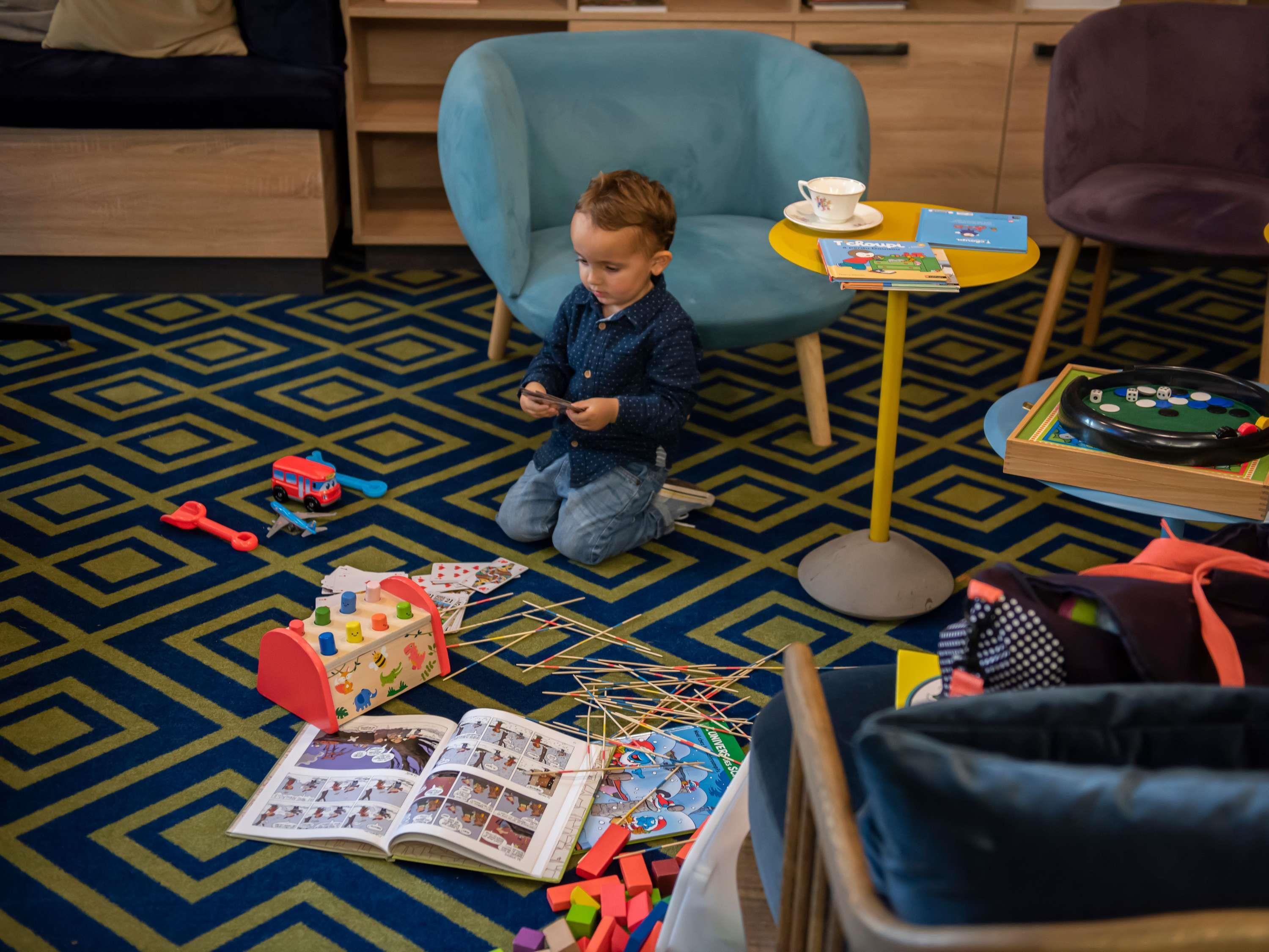 A child playing with toys on a patterned carpet, a small table with a cup, and a book on the floor.