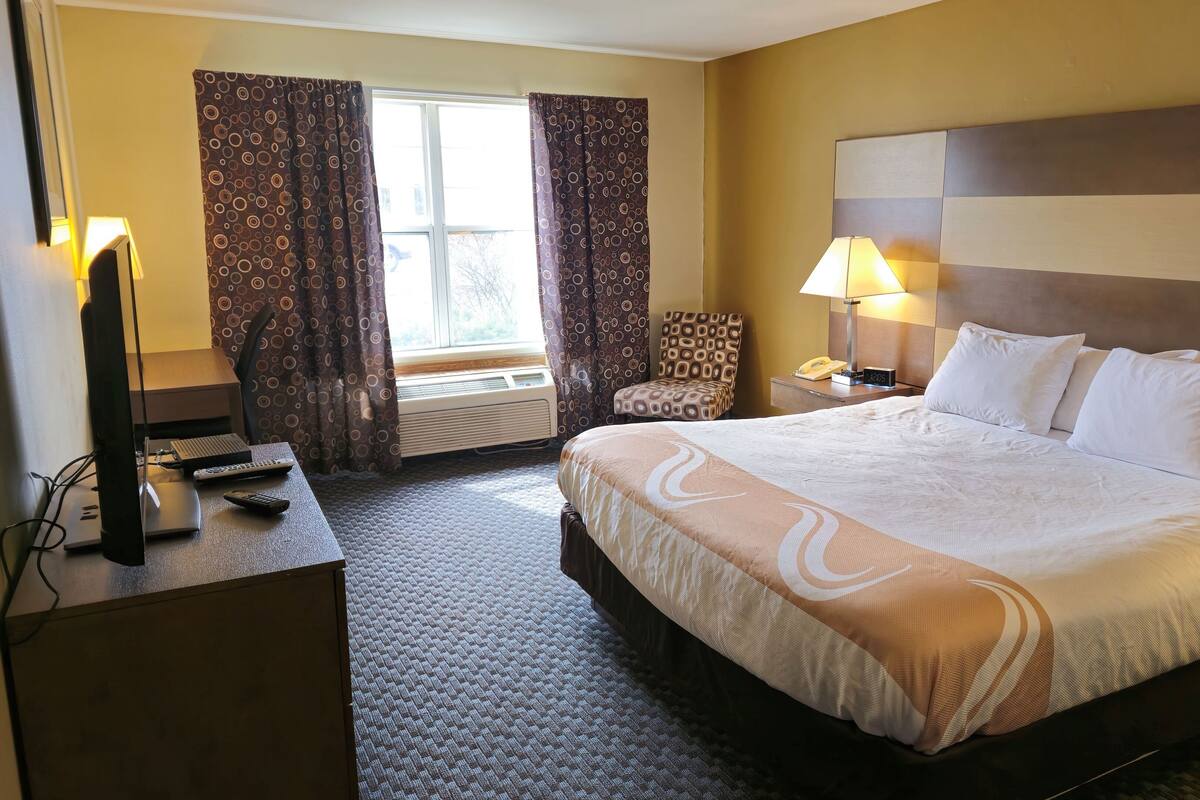 A hotel room with a large bed, a desk with a computer, a chair, and a window with curtains.