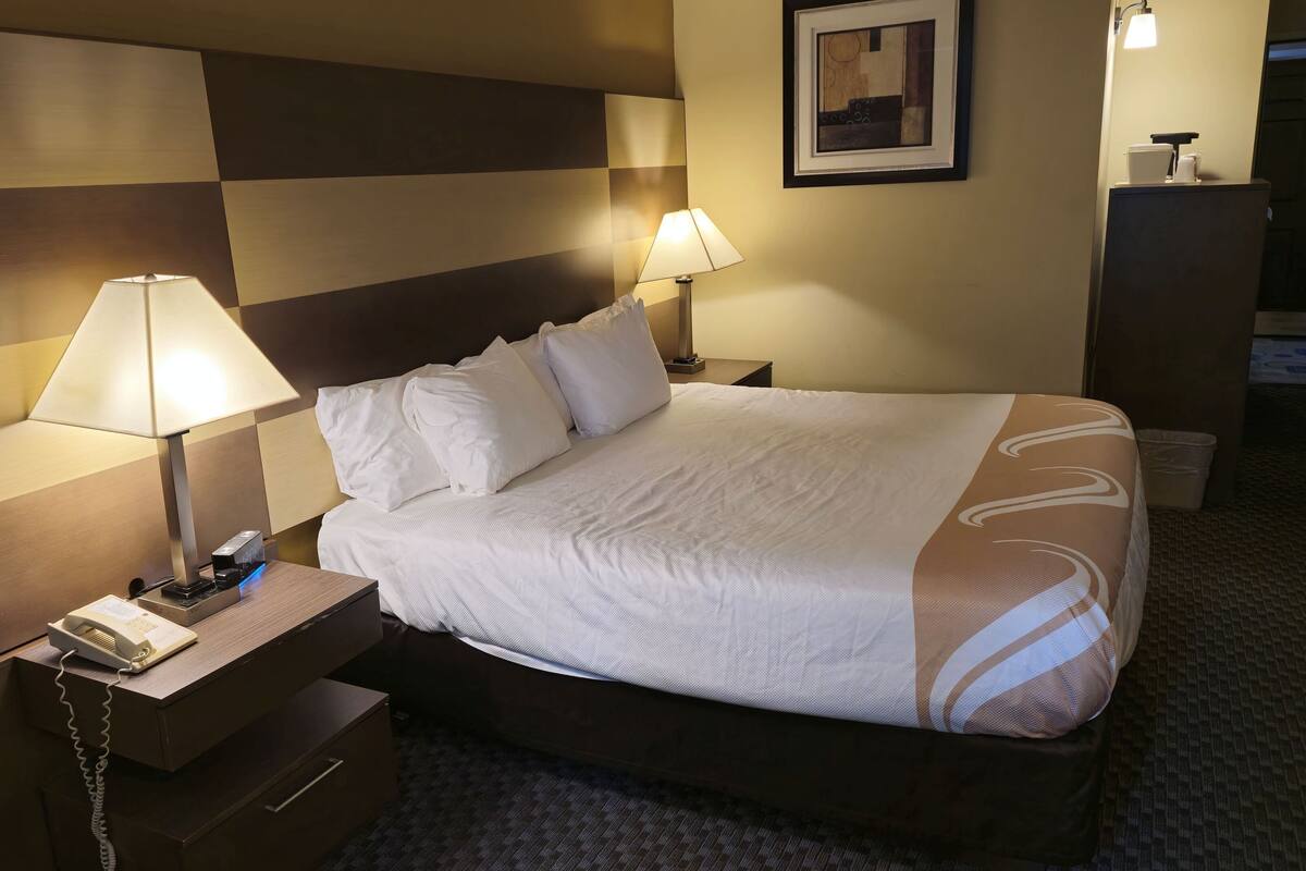 A hotel room with a bed, two bedside lamps, a nightstand with a telephone, and a framed picture on the wall.