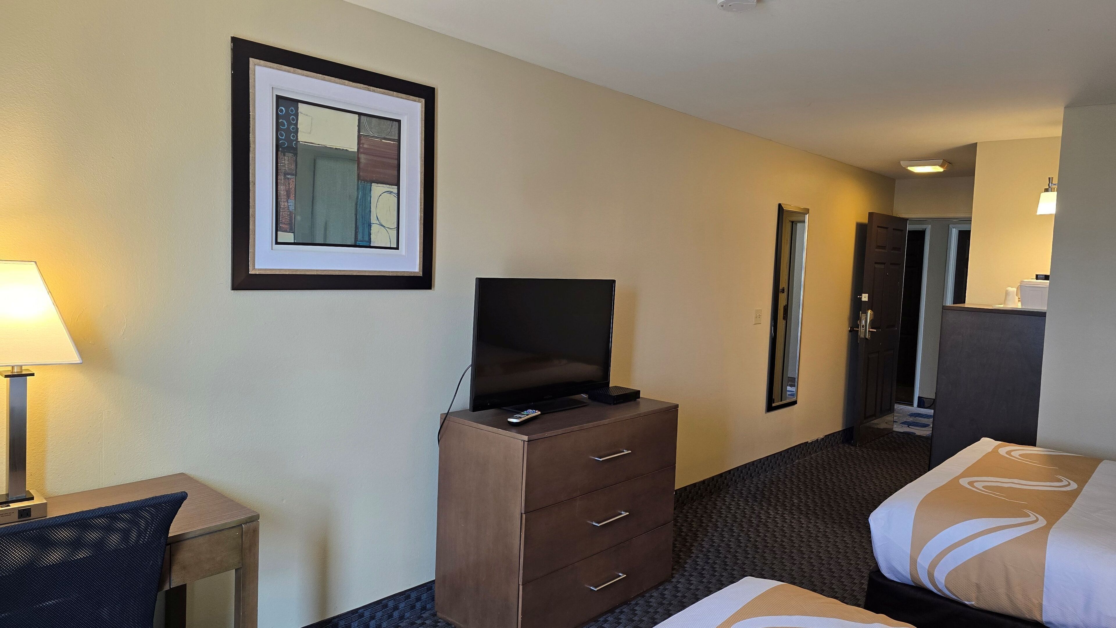 A hotel room with a bed, a desk with a lamp, a TV on a dresser, and a framed picture on the wall.